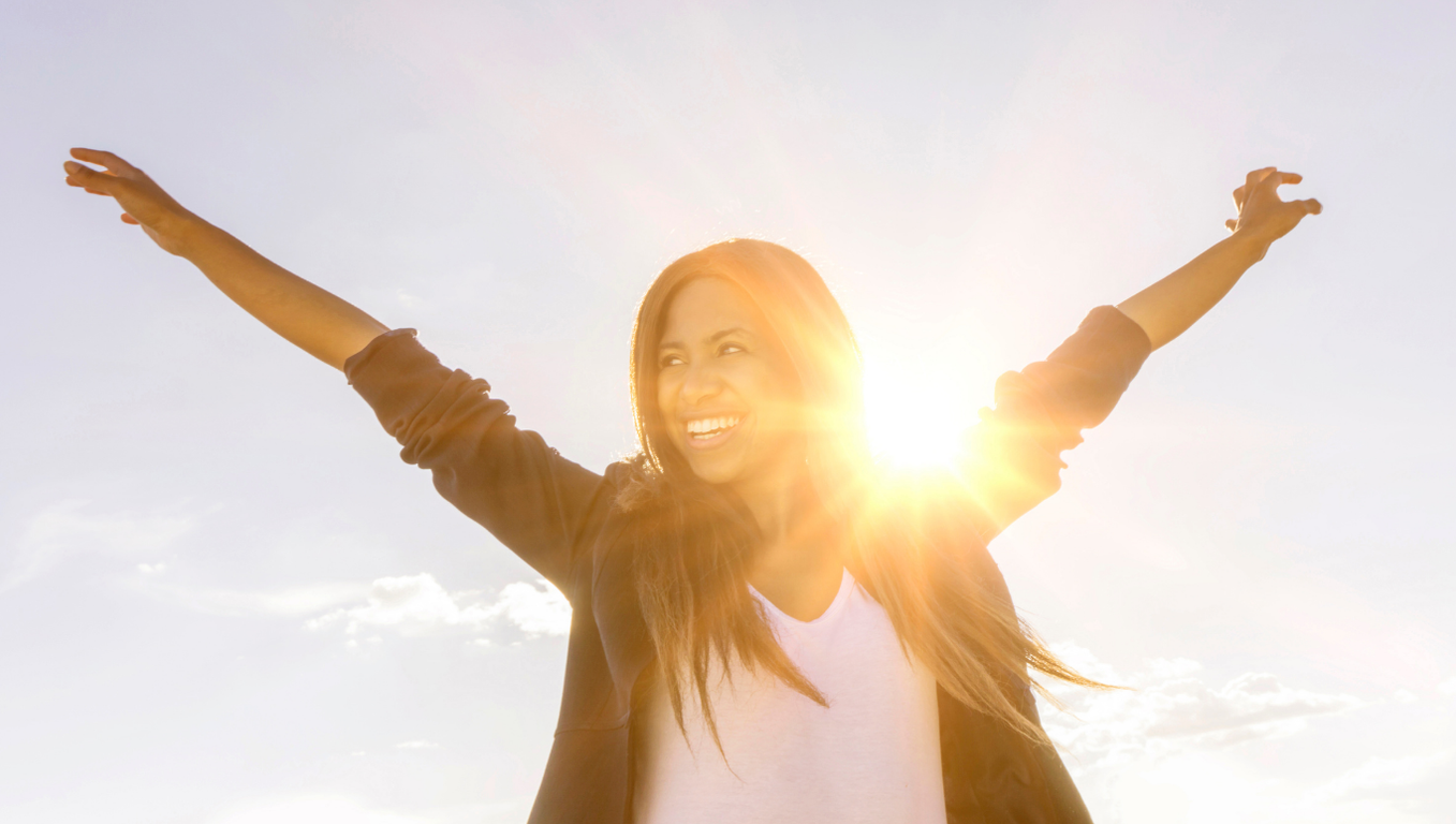 Woman, backlit by rays of sunshine, raising her arms in celebration of her ketamine therapy success